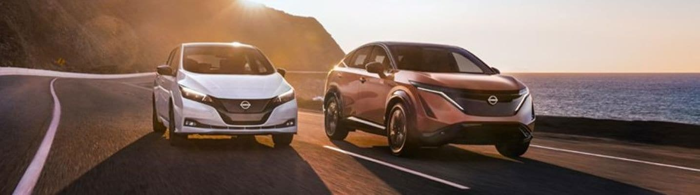 Two Nissan EVs, a Leaf and Ariya, driving on a coastal road at sunset.